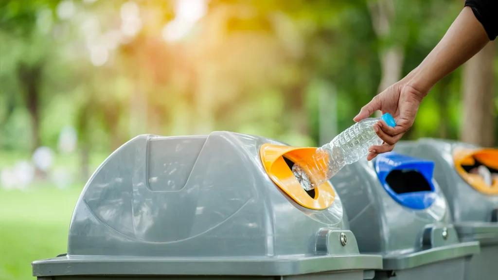 Mano che getta una bottiglia di plastica in un bidone della raccolta differenziata all'aperto in un parco.
